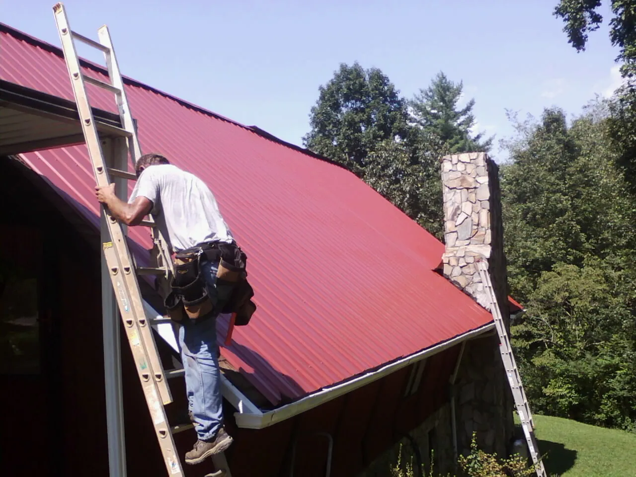 Roofer on a ladder beside a red metal roof and stone chimney — Soffit & Fascia Repair by Roofing Brothers in Elkridge